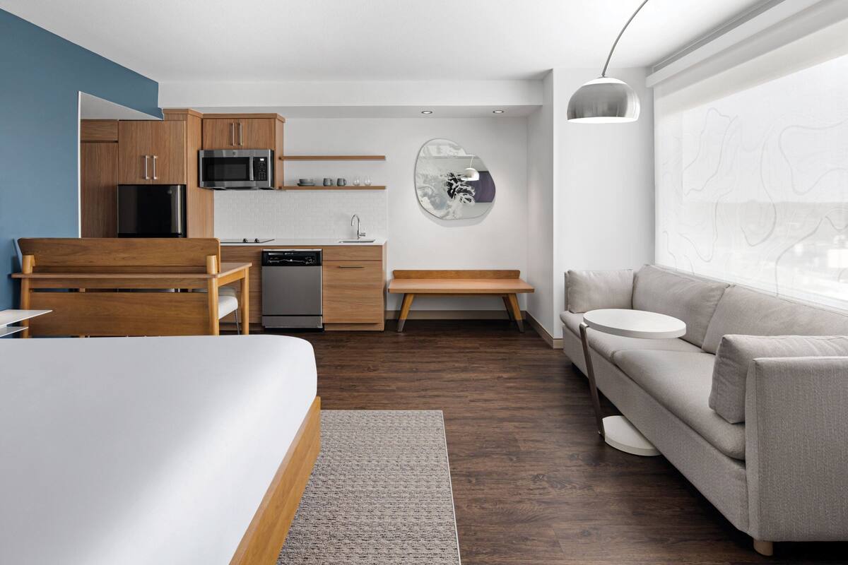 A modern hotel room with a bed, a sofa, a dining area with a table and chairs, a kitchen with a microwave and refrigerator, and a large window with a view.