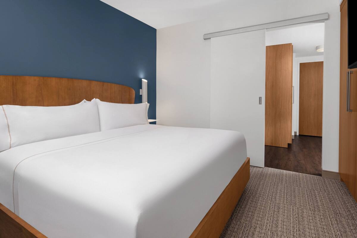 A hotel room with a large bed, a wooden headboard, a white wall, and a door leading to a hallway.