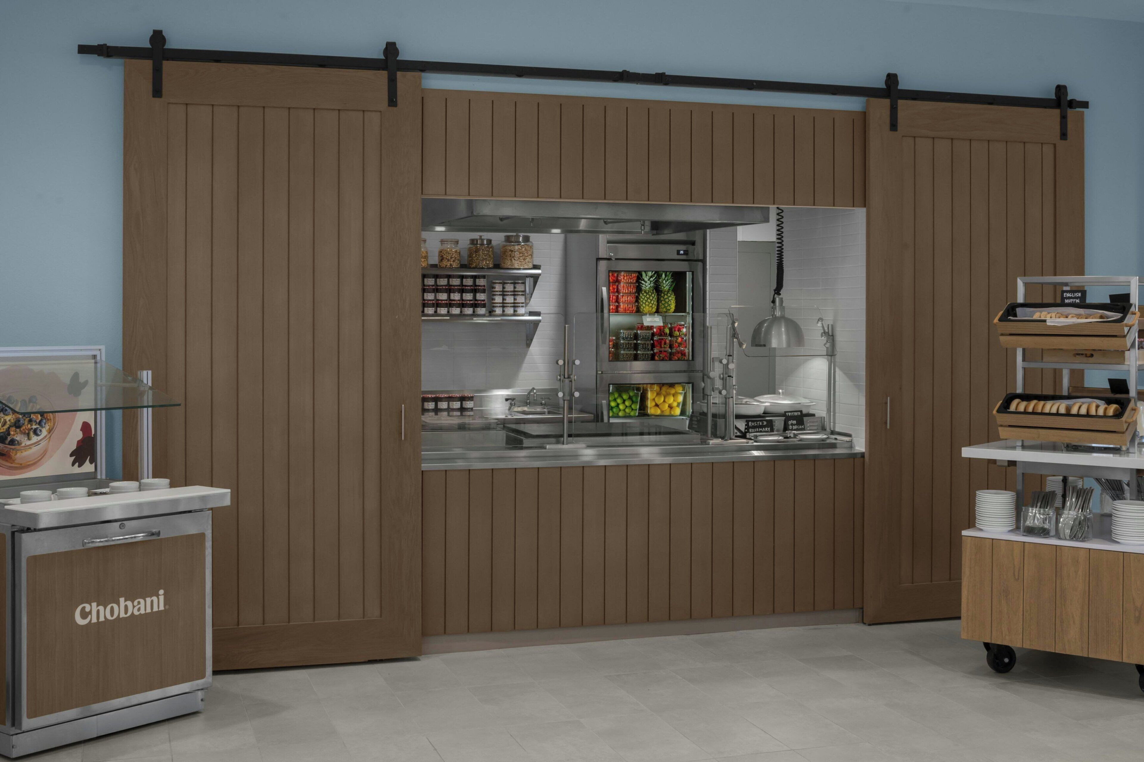 Una kitchenette con un refrigerador Chobani, una cafetera y un expositor de alimentos con diversos productos.