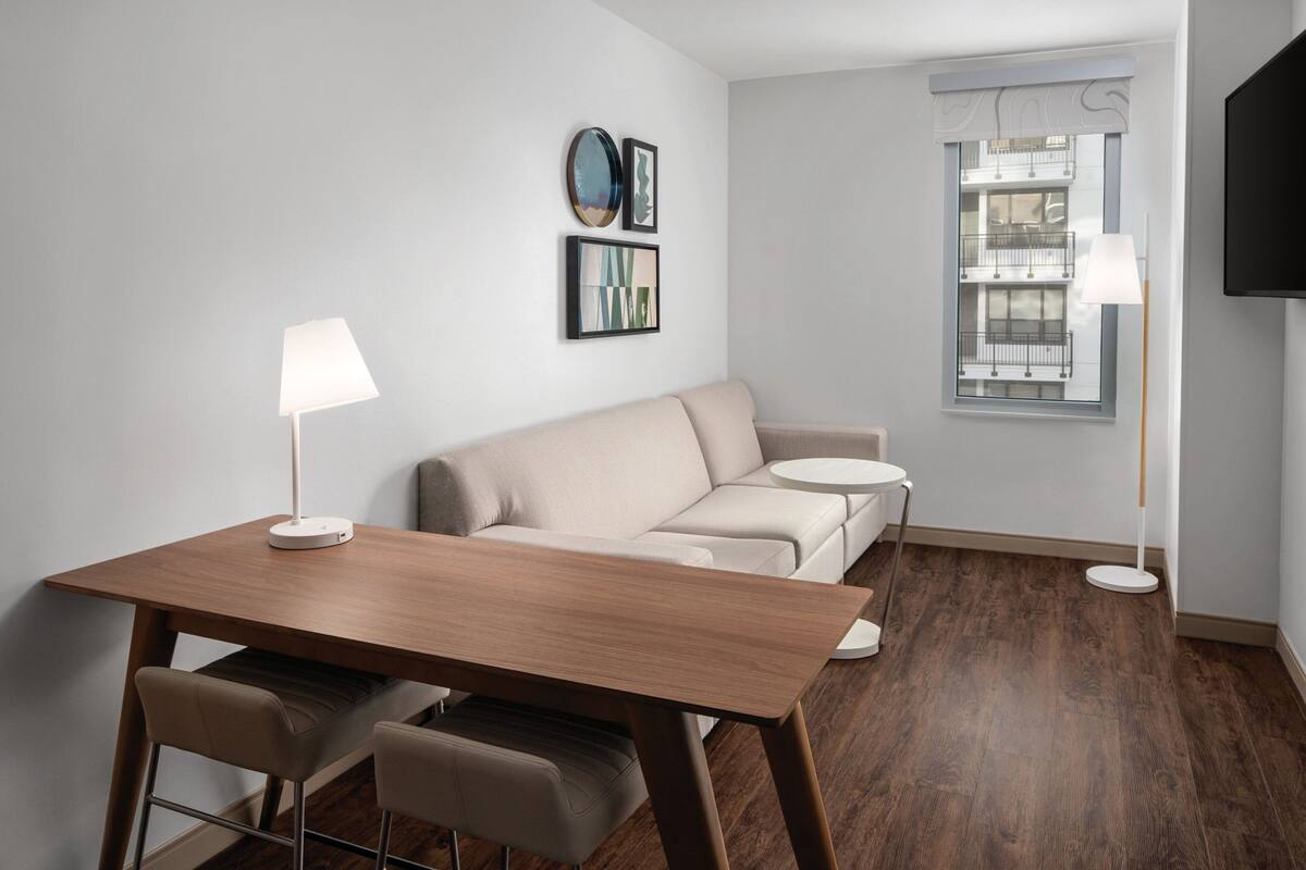 A modern living room with a wooden dining table, two chairs, a beige sofa, a small round table, and a window with a view of buildings.