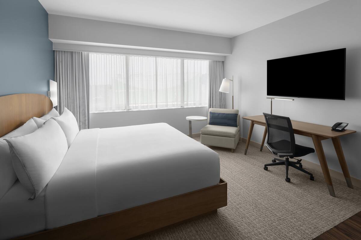 A hotel room with a large bed, a desk with a chair, a TV, and a window with curtains.