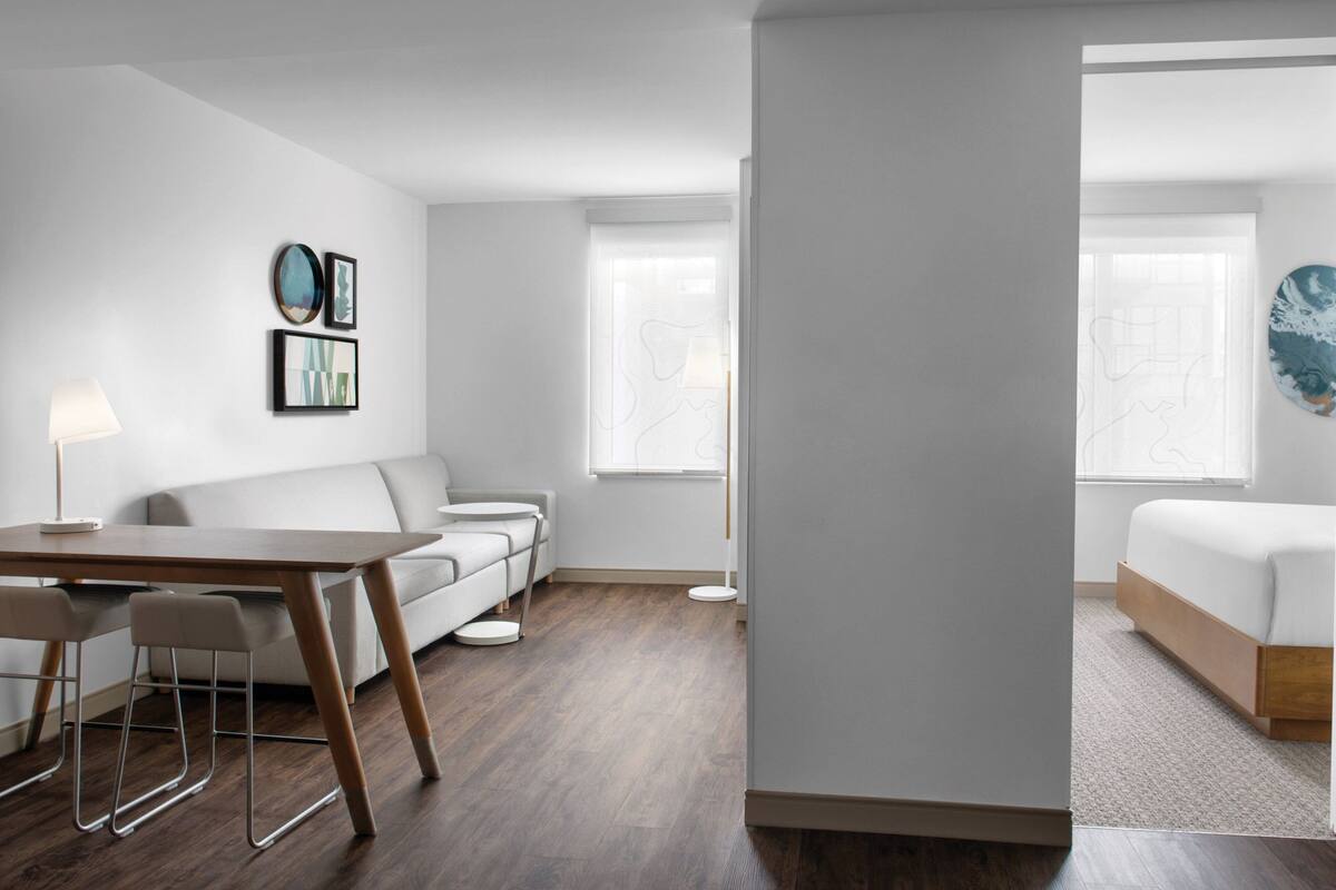 A modern, minimalist living room with a wooden dining table, two white sofas, and a large window providing natural light.