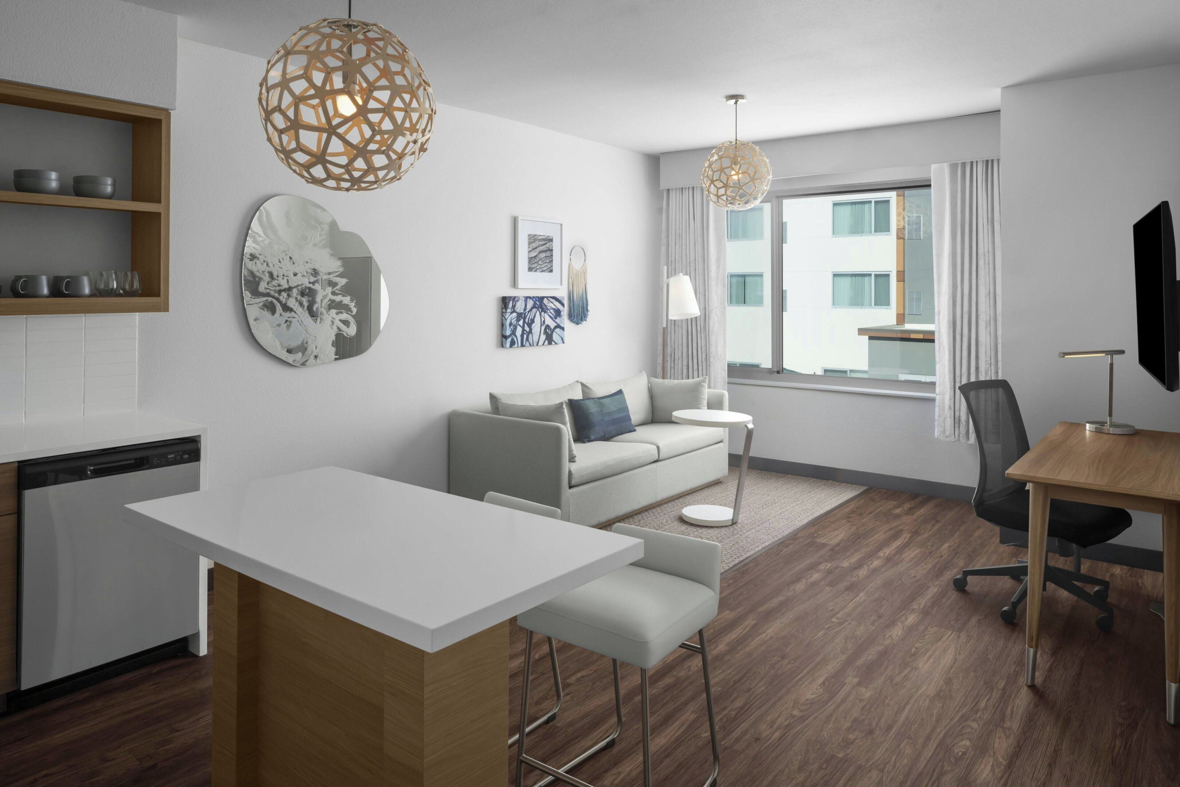 A modern hotel room with a kitchenette, a dining area, a sofa, a TV, and a large window with curtains.
