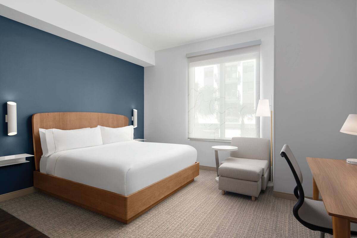 A modern hotel room with a large bed, a small armchair, a round table, and a window with a view.