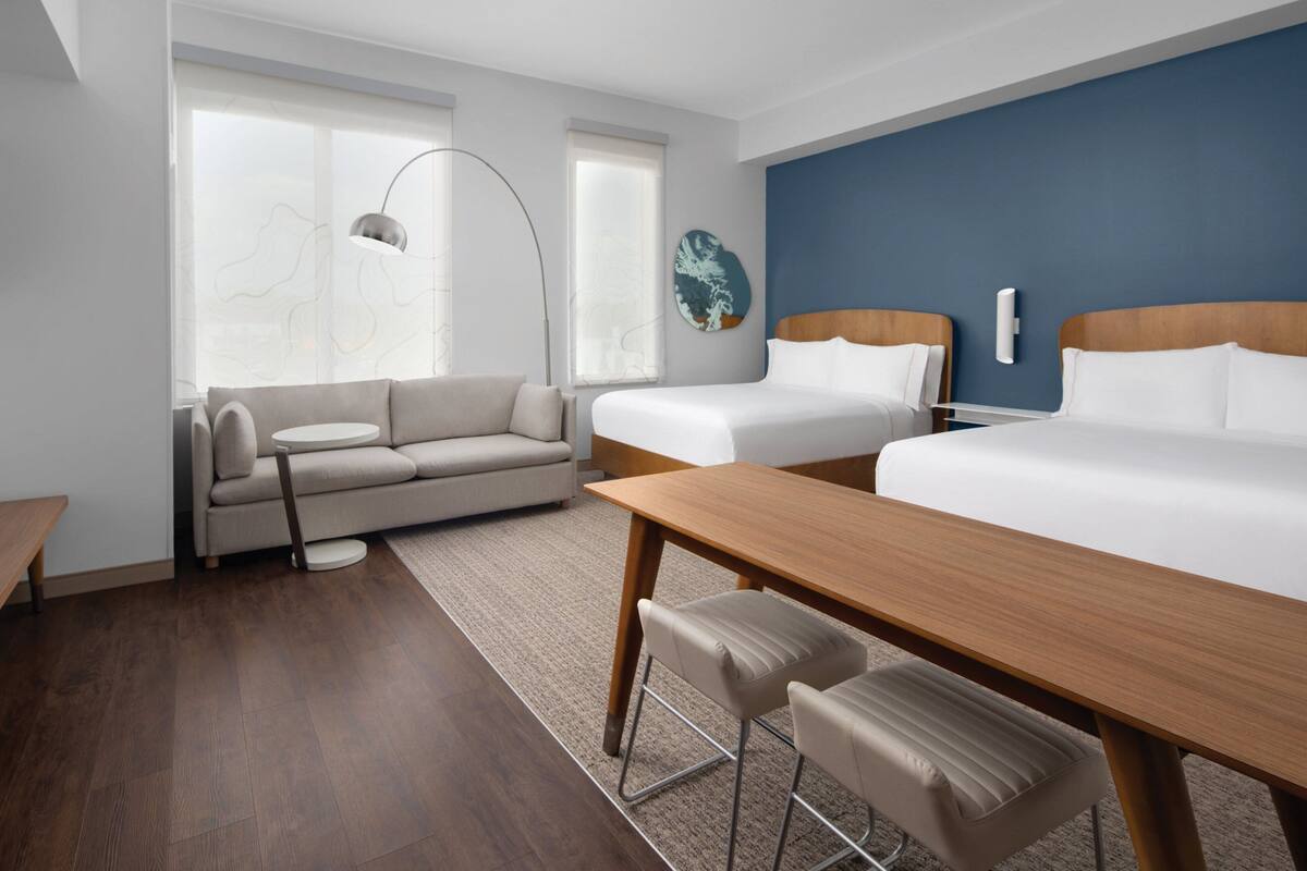 A modern hotel room with a wooden dining table, two single beds, a sofa, and a large window with a view.