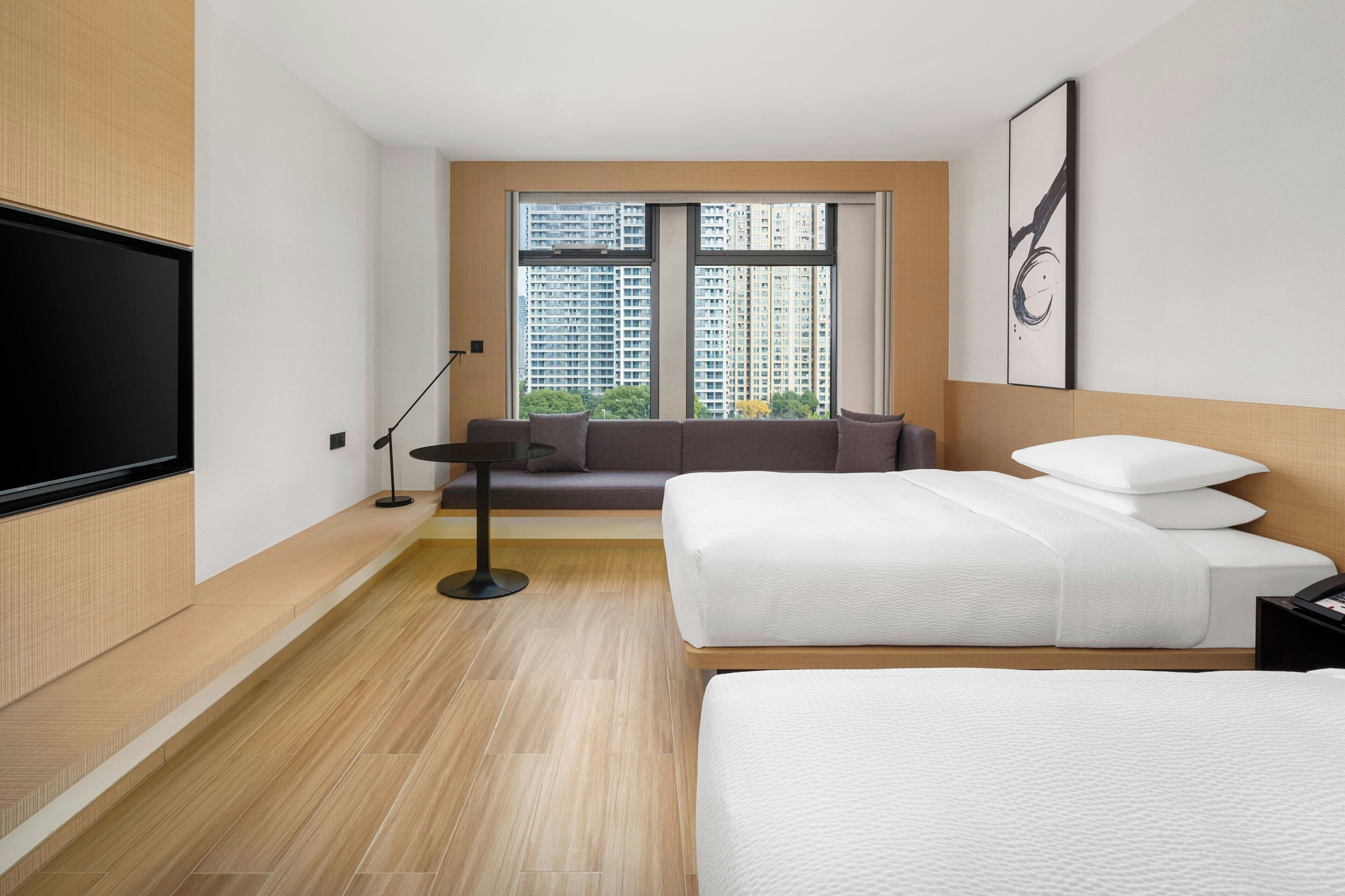 Ein modernes Hotelzimmer mit einem großen Bett, einer Couch, einem Fernseher und einem Fenster mit Blick auf die Stadt.