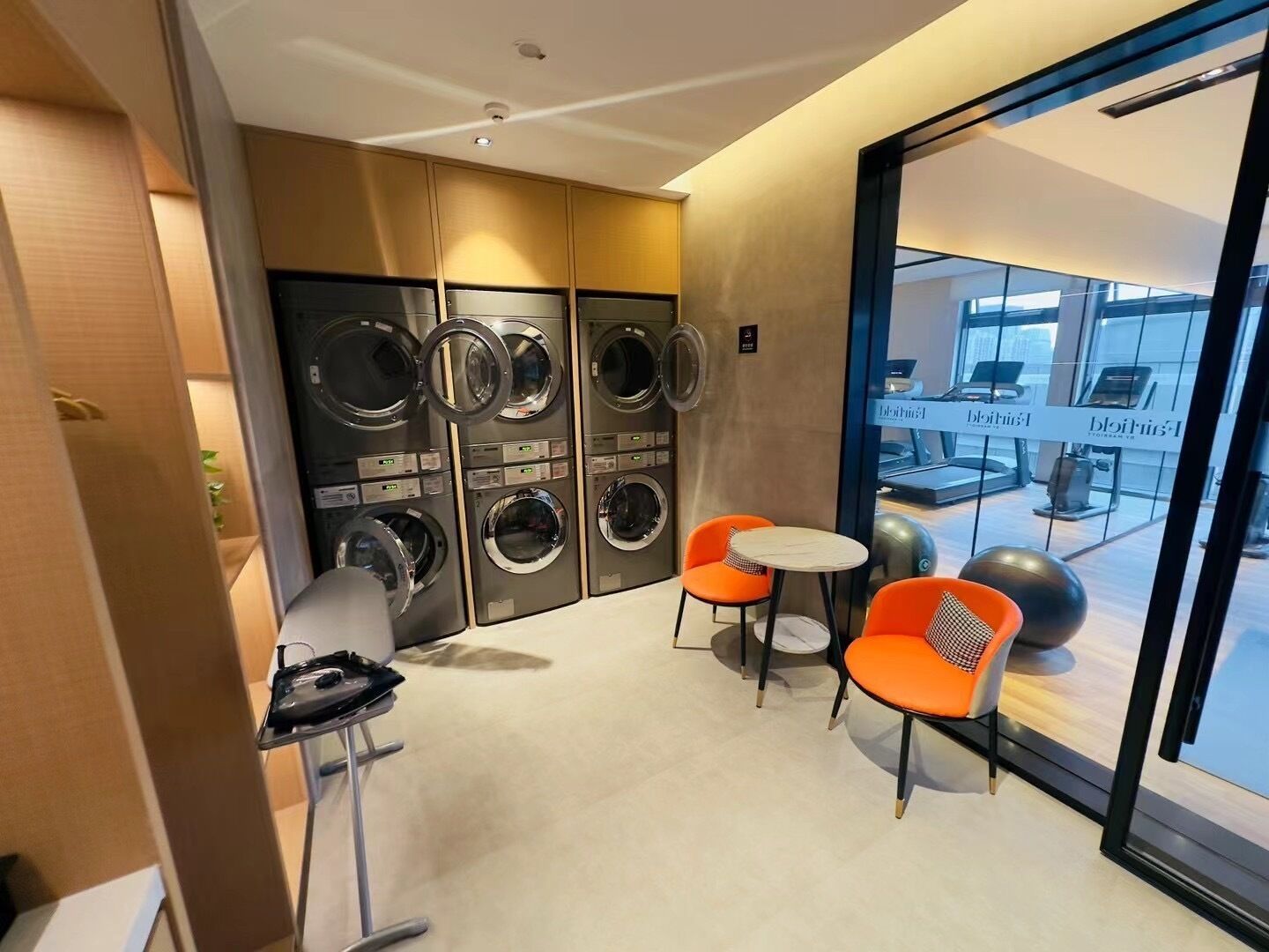 A hotel room with a washing machine, two orange chairs, a small table, and a view of the gym area.