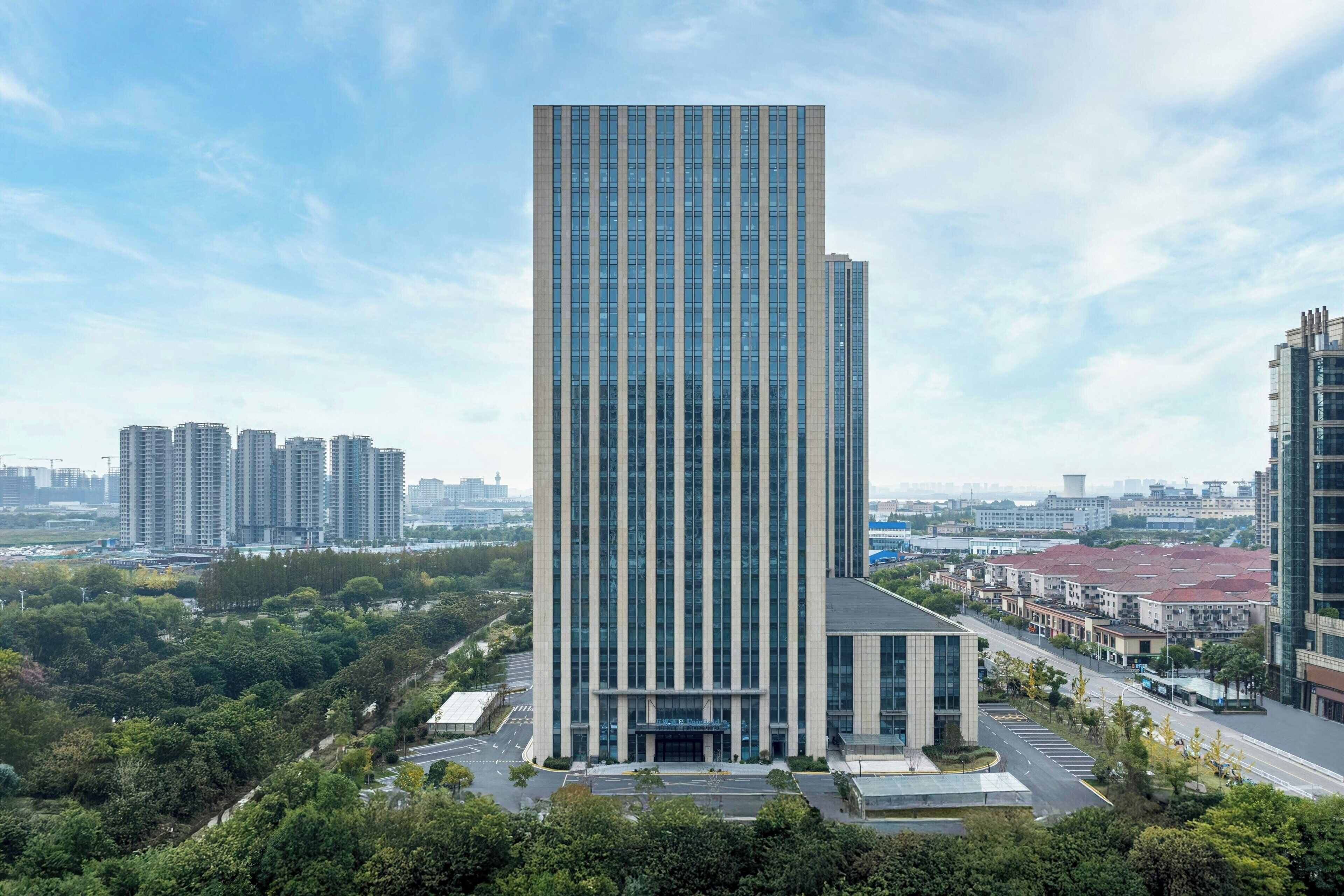 A tall, modern skyscraper with a glass facade, surrounded by greenery and other buildings.