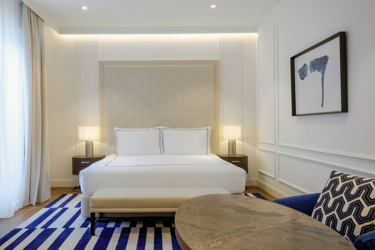 A modern hotel room with a large bed, two bedside tables, a marble side table, and a patterned throw pillow.