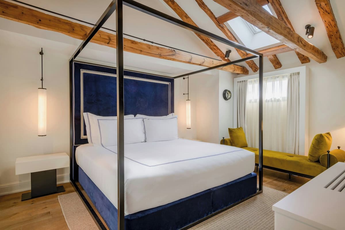 A four-poster bed with a dark headboard, a bedside table, a sofa, and a view of the outdoors through a window with curtains.
