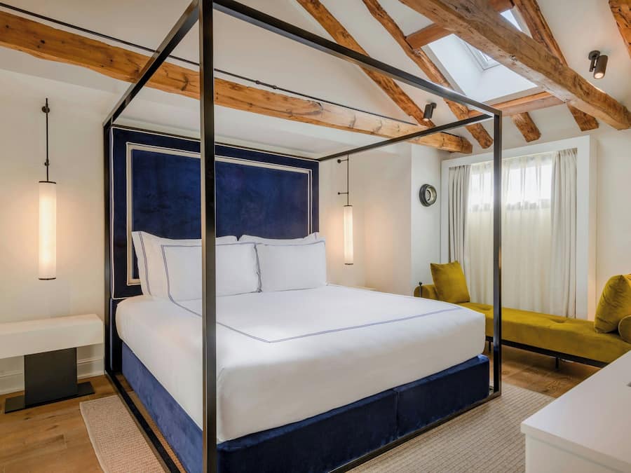 Una cama con dosel, un cabecero oscuro, una mesita de noche, un sofá y vistas al exterior a través de una ventana con cortinas.