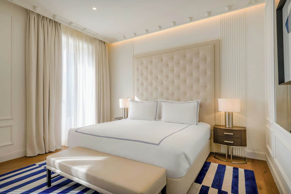 A hotel room with a large bed, a bedside table with a lamp, a bench, and a striped rug.