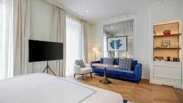 A hotel room with a large bed, a flat-screen TV, a blue sofa, a small white armchair, a round coffee table, and a bookshelf with decorative items.