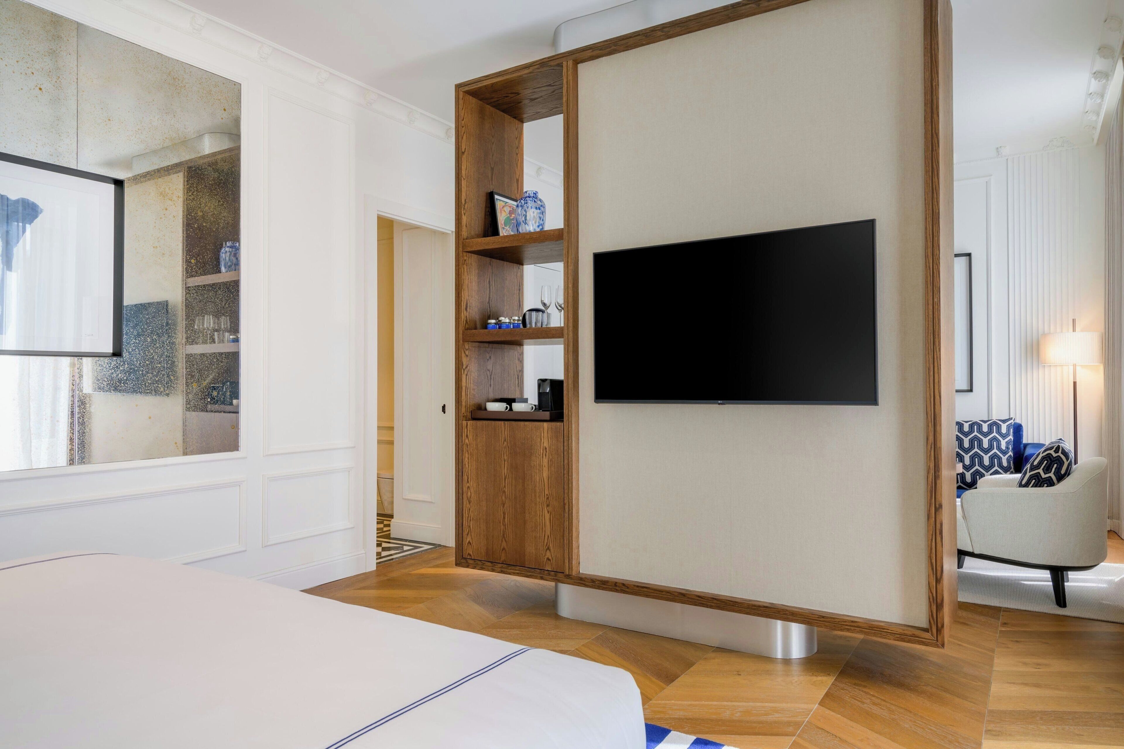 Une chambre d’hôtel moderne avec un grand lit, un téléviseur fixé au mur, un meuble en bois avec des étagères et un coin salon comprenant une lampe et un fauteuil.
