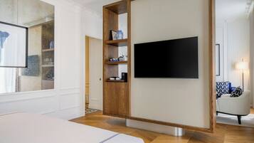 Une chambre d’hôtel moderne avec un grand lit, un téléviseur fixé au mur, un meuble en bois avec des étagères et un coin salon comprenant une lampe et un fauteuil.