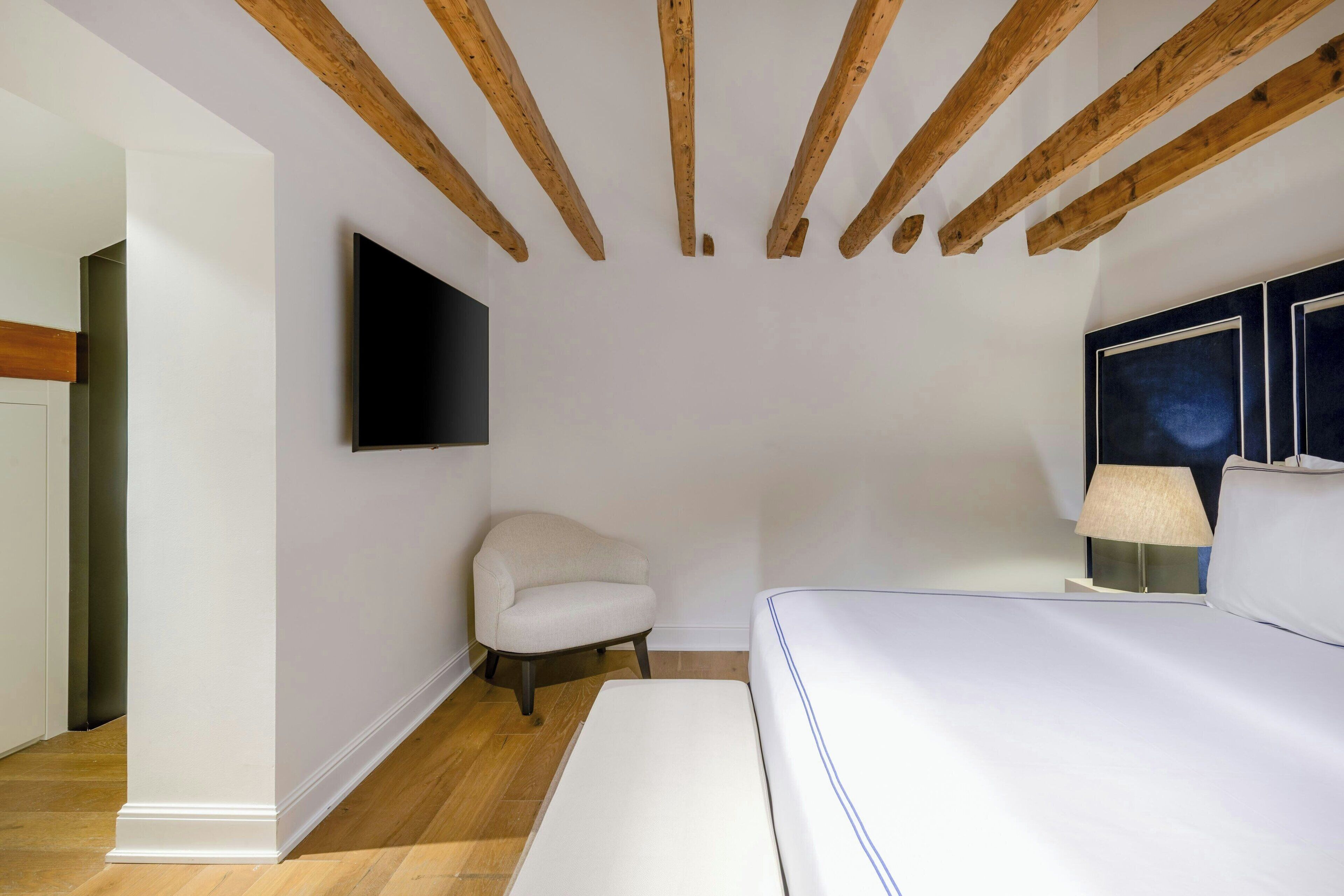 A bedroom with a wooden ceiling, a flat-screen TV mounted on the wall, a white sofa, and a bedside table with a lamp.