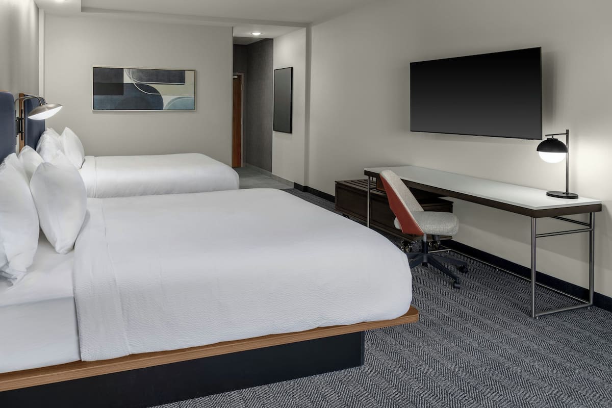 A hotel room with two beds, a desk, a chair, a TV, and artwork on the walls.
