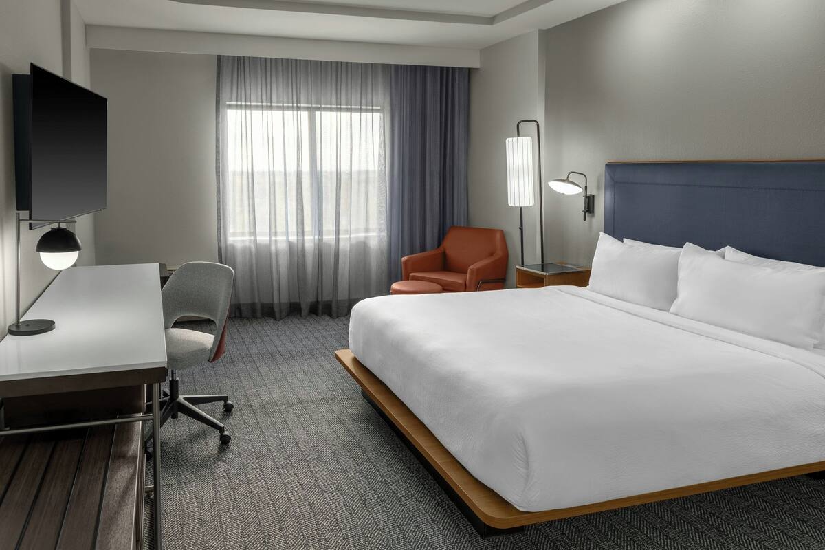 A hotel room with a large bed, a desk with a chair, a TV, and a window with curtains.