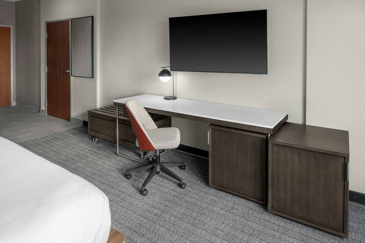 A modern hotel room with a large bed, a desk with a chair, a flat-screen TV, and a wardrobe.