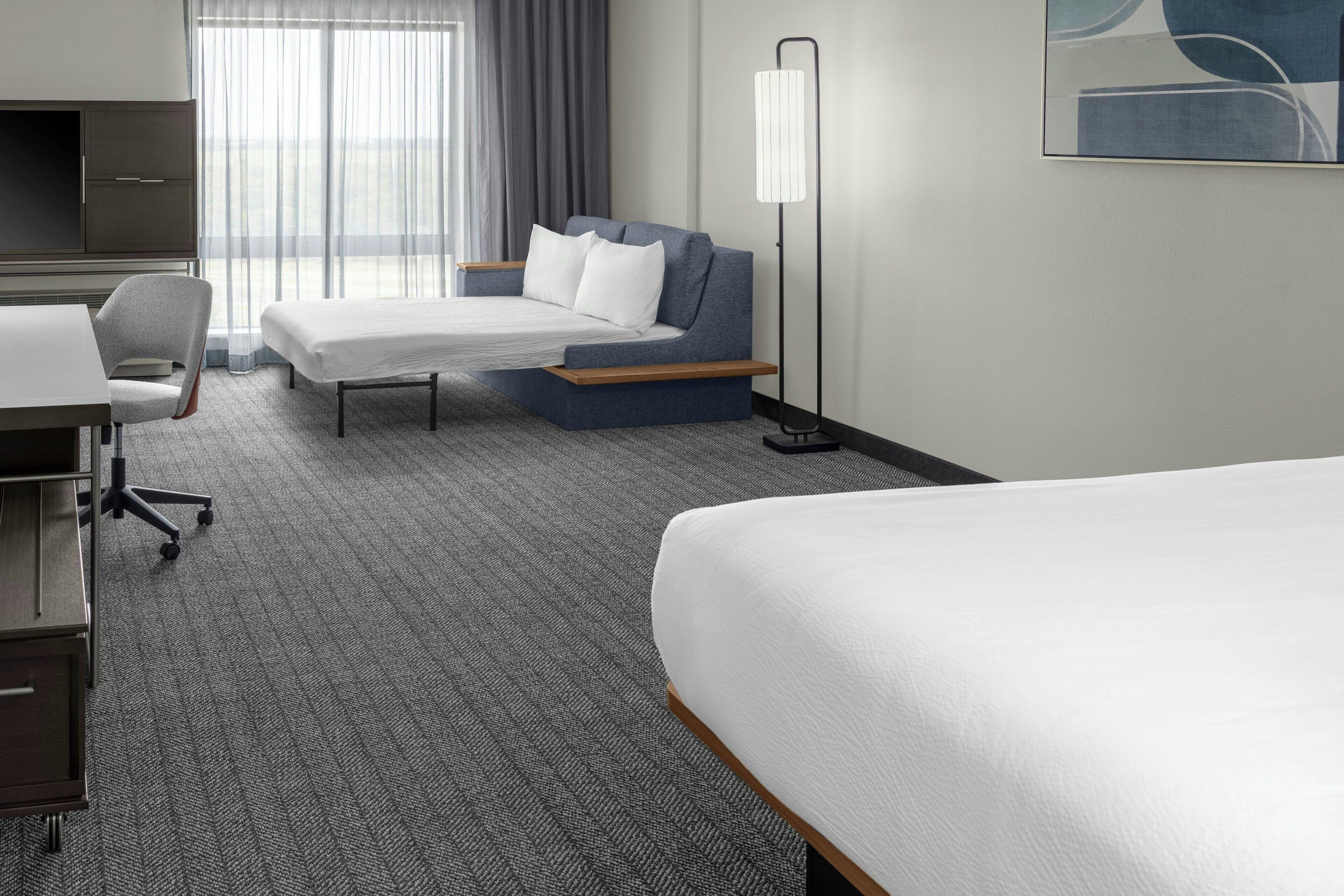 Una habitación de hotel con cama, escritorio, silla, mesita de noche y lámpara de pie.