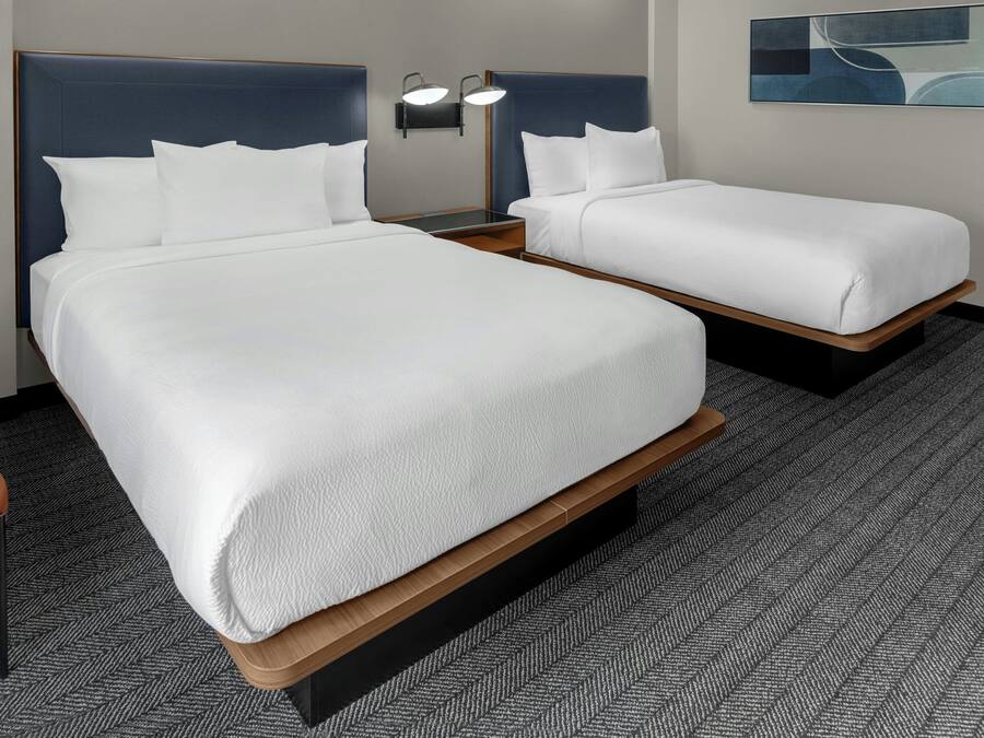 Habitación de hotel con dos camas, una mesita de noche con lámpara, una silla y suelo cubierto de alfombra.