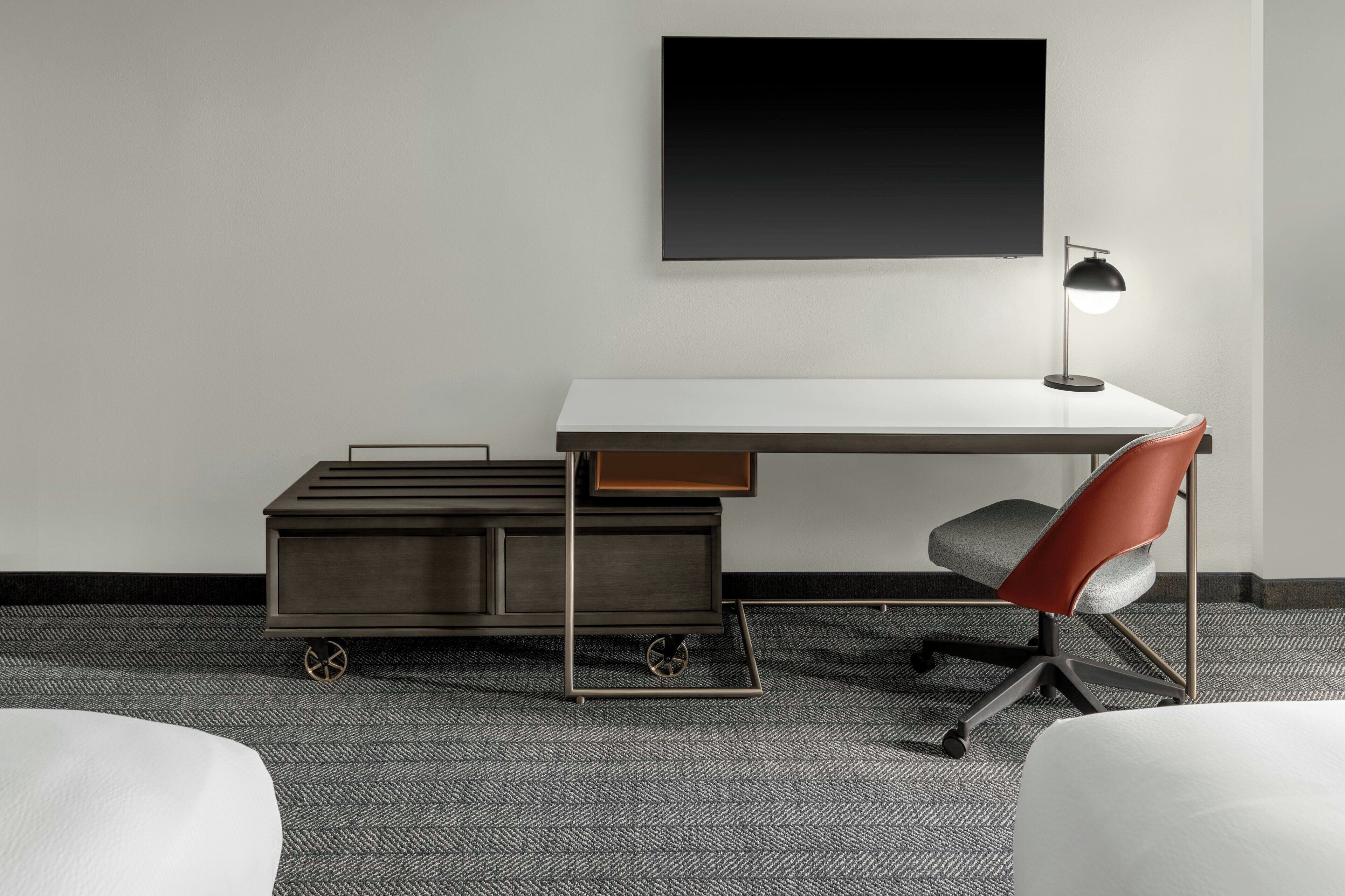 A modern hotel room with a white desk, a red office chair, a black lamp, and a grey carpet.