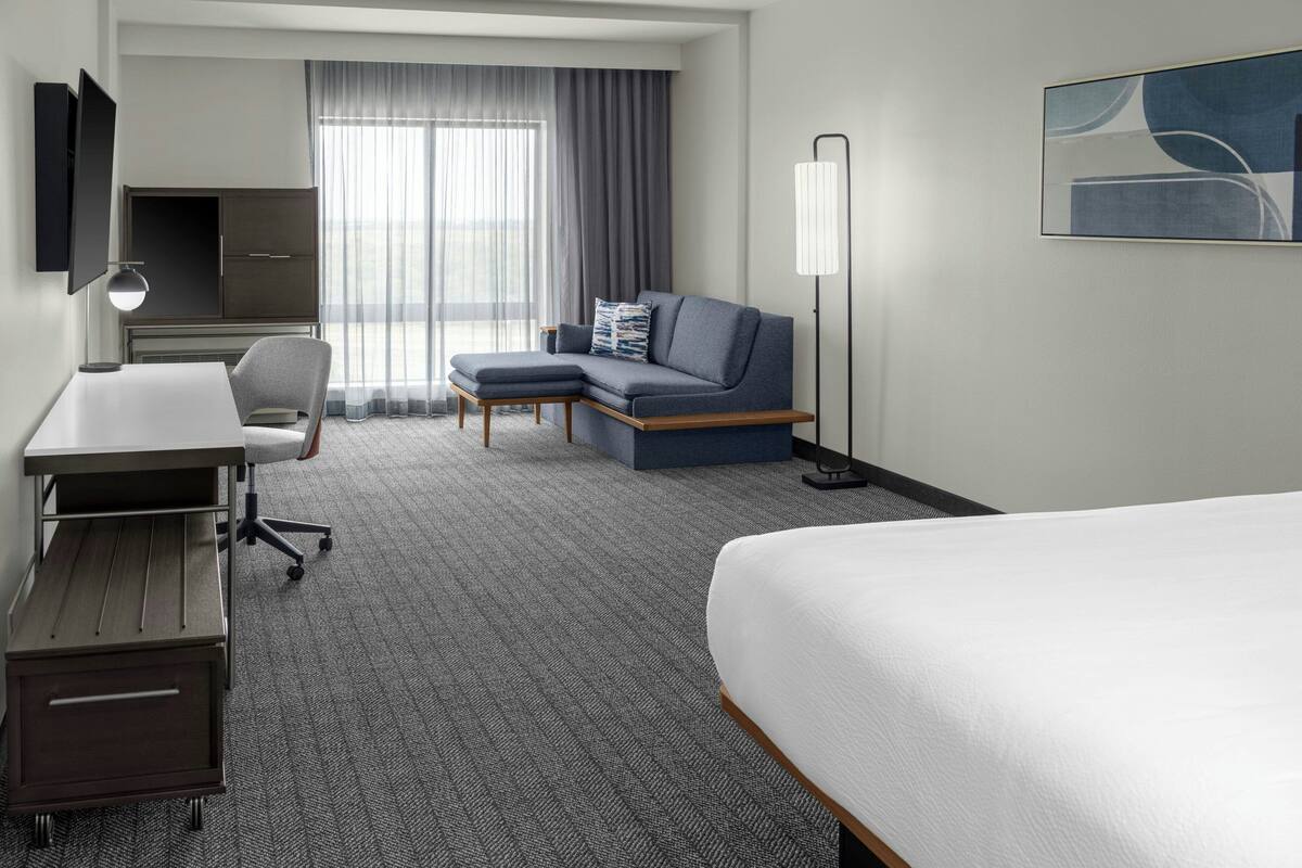 A hotel room with a bed, a desk, a chair, a sofa, a TV, and a lamp.