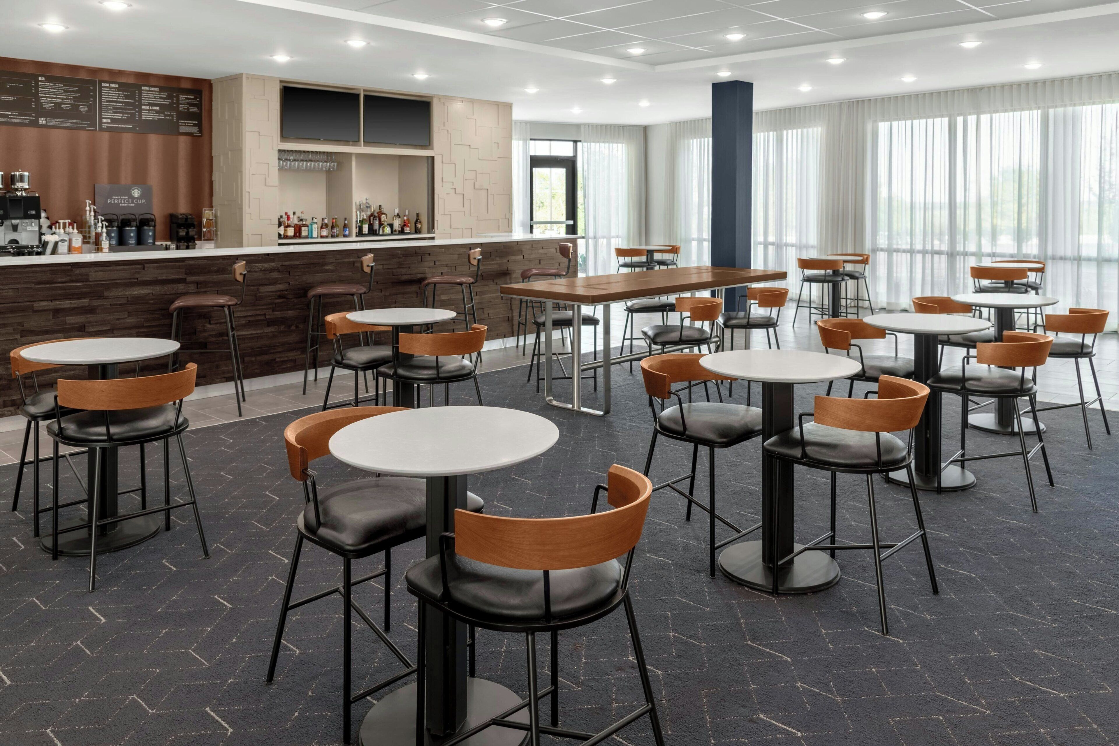 A modern restaurant with round tables and chairs, a bar area, and large windows.