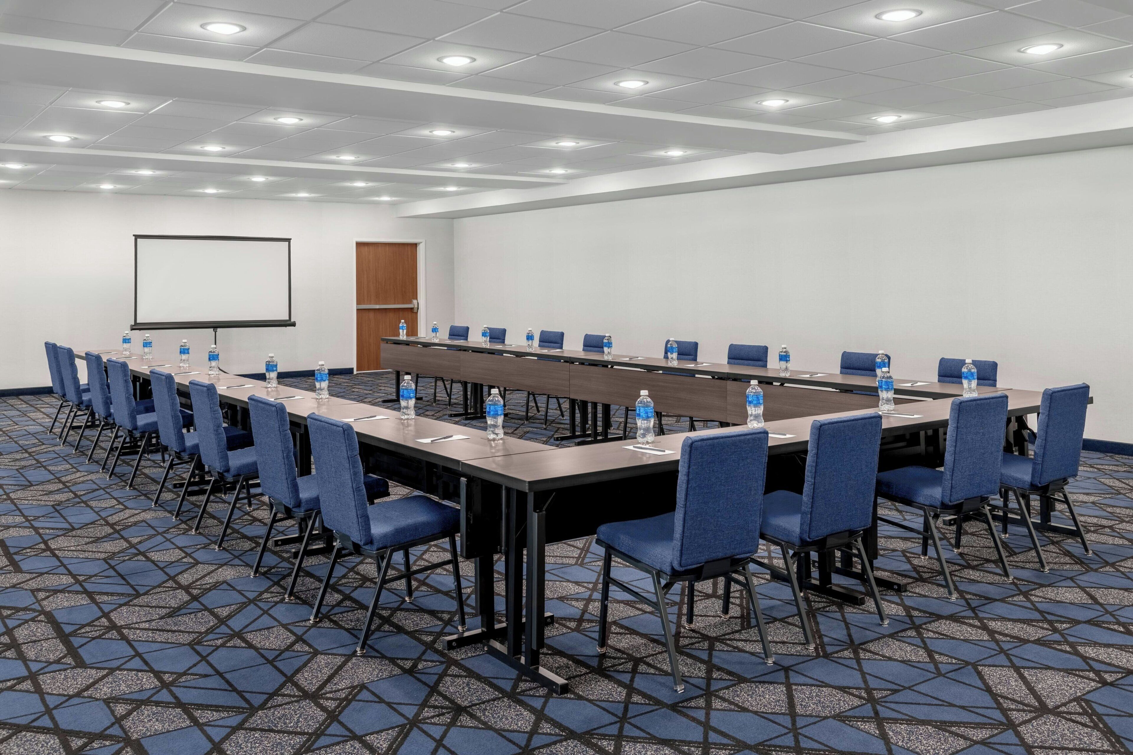 Un salón de conferencias con una mesa larga, sillas azules, botellas de agua, una pantalla de proyector y suelo alfombrado.