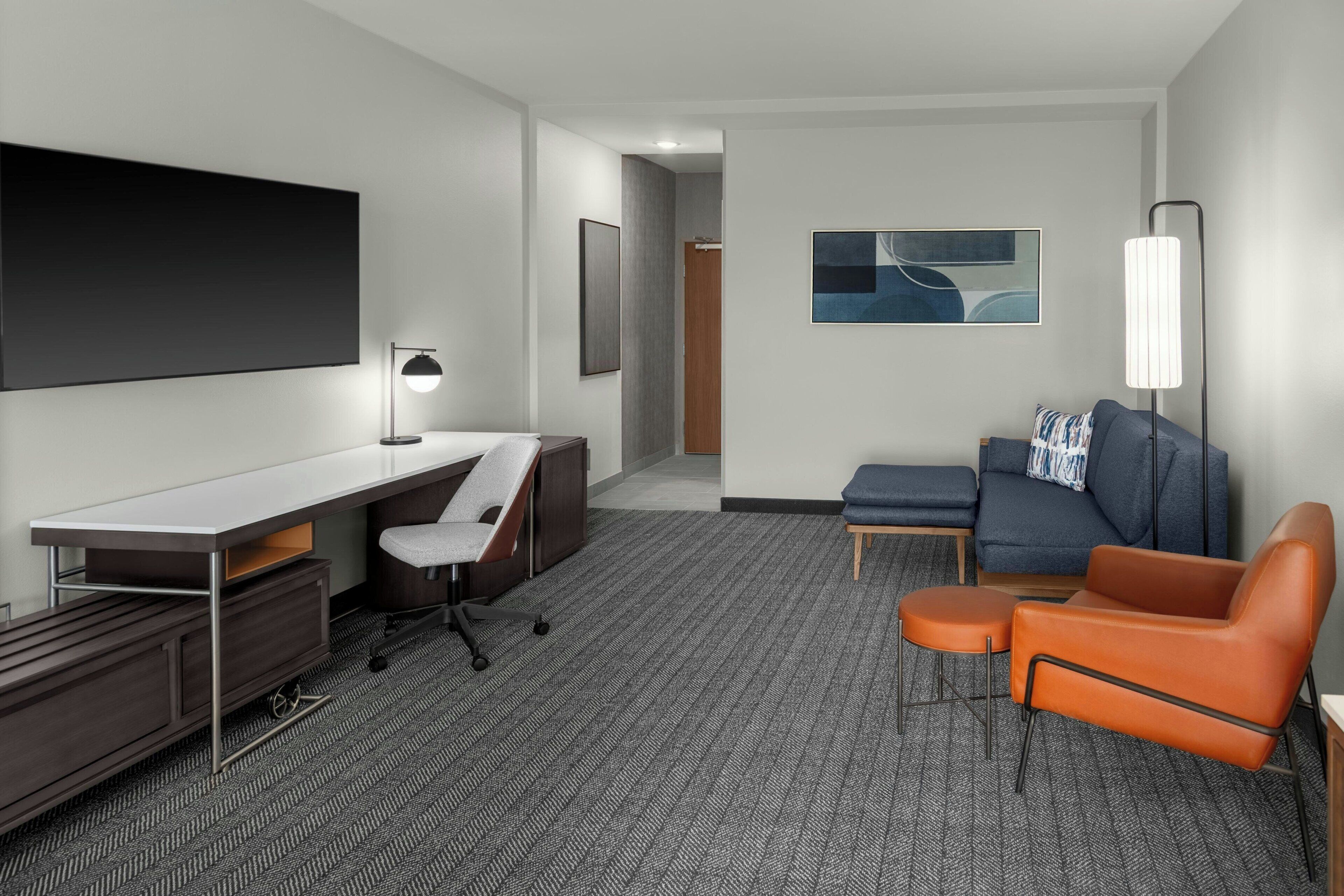 Une chambre d’hôtel moderne dotée d’une grande télévision à écran plat, d’un bureau avec une chaise, d’un canapé et d’un petit pouf.