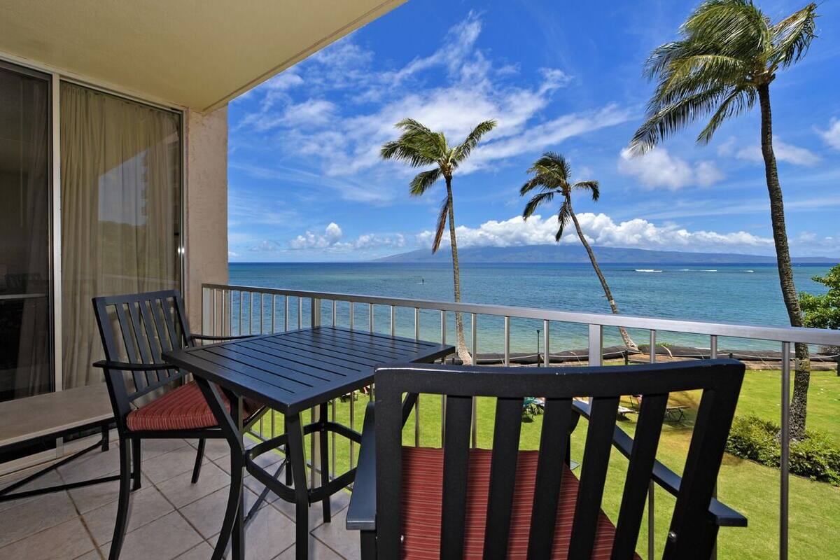 Ein Balkon mit Tisch und Stühlen, der Blick auf einen Strand mit Palmen und einem klaren Himmel bietet.