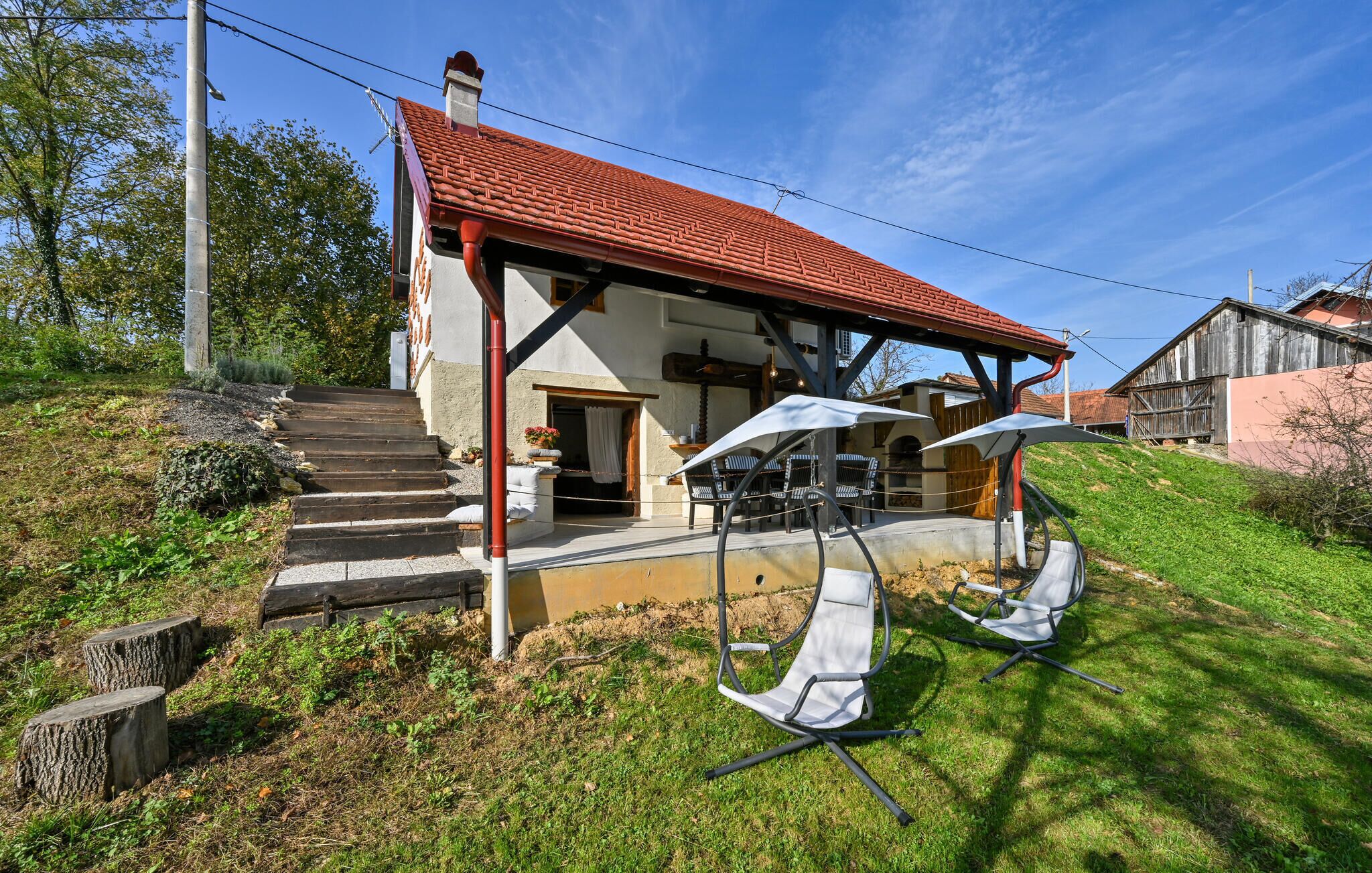 Uma casa com telhado de telhas vermelhas, um alpendre com móveis de exterior e uma escadaria de pedra que leva à entrada.