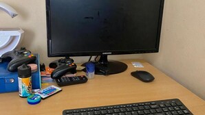 Un escritorio con un monitor Samsung, teclado, ratón, un control de videojuegos y varios objetos como una botella, un mando a distancia y una tarjeta pequeña.