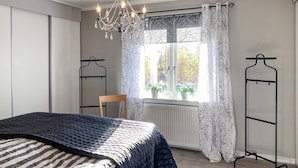 Un dormitorio con cama, una silla, un ventanal con cortinas, un radiador y una araña.