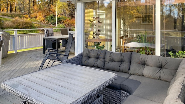 Un área de estar exterior techada con sofá acolchado y mesa, rodeada por una terraza de madera y paredes de vidrio que ofrecen vistas a un área boscosa.