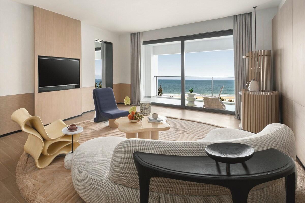 A modern hotel room with a large TV, comfortable seating, a small table, and a view of the ocean.