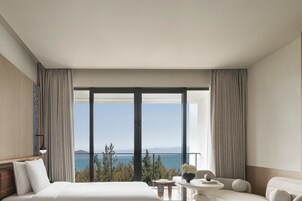 Una habitación de hotel con una cama grande, una mesita con un florero, un sofá y vista al mar a través de ventanales del suelo al techo.