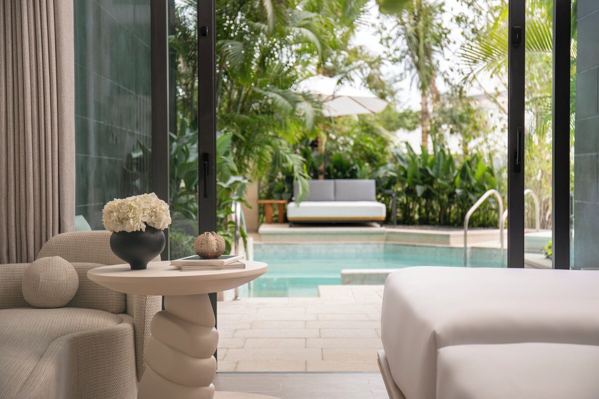 A modern hotel room with a pool view, a sofa, a round table with a vase, and a bed.