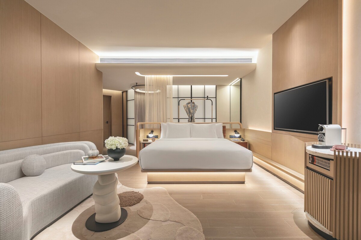 Una habitación de hotel moderna con una cama grande, un sofá, una mesita con un florero y un televisor montado en la pared.