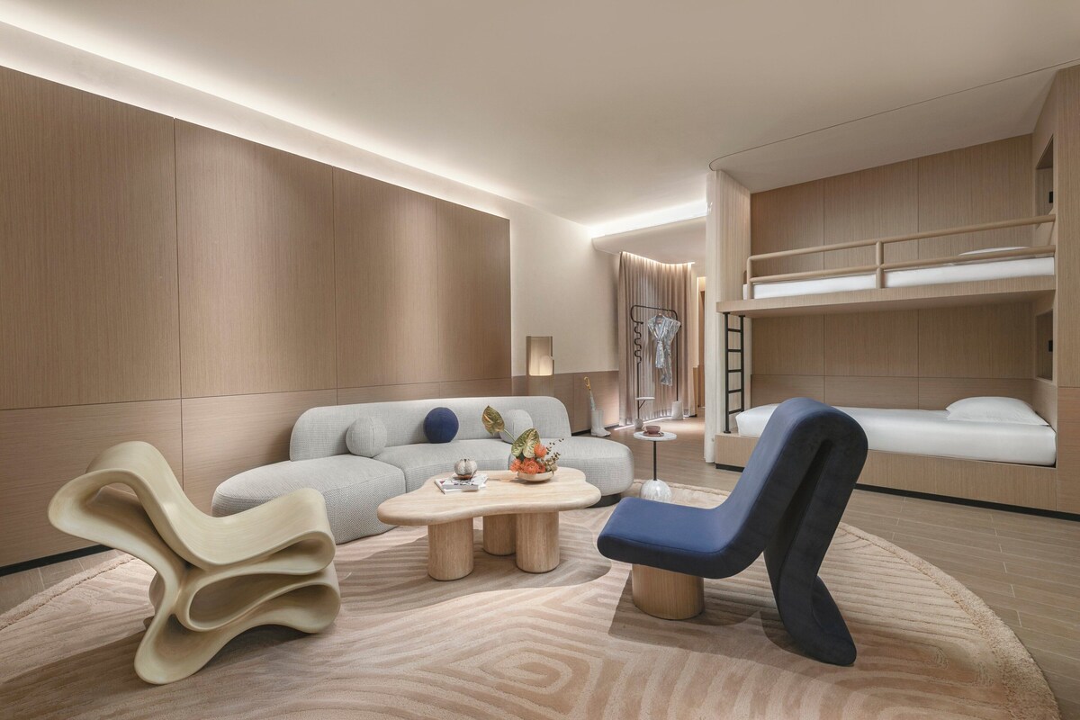 Una habitación de hotel moderna con sofá, una silla original, una mesita de café pequeña y una cama individual sobre otra.