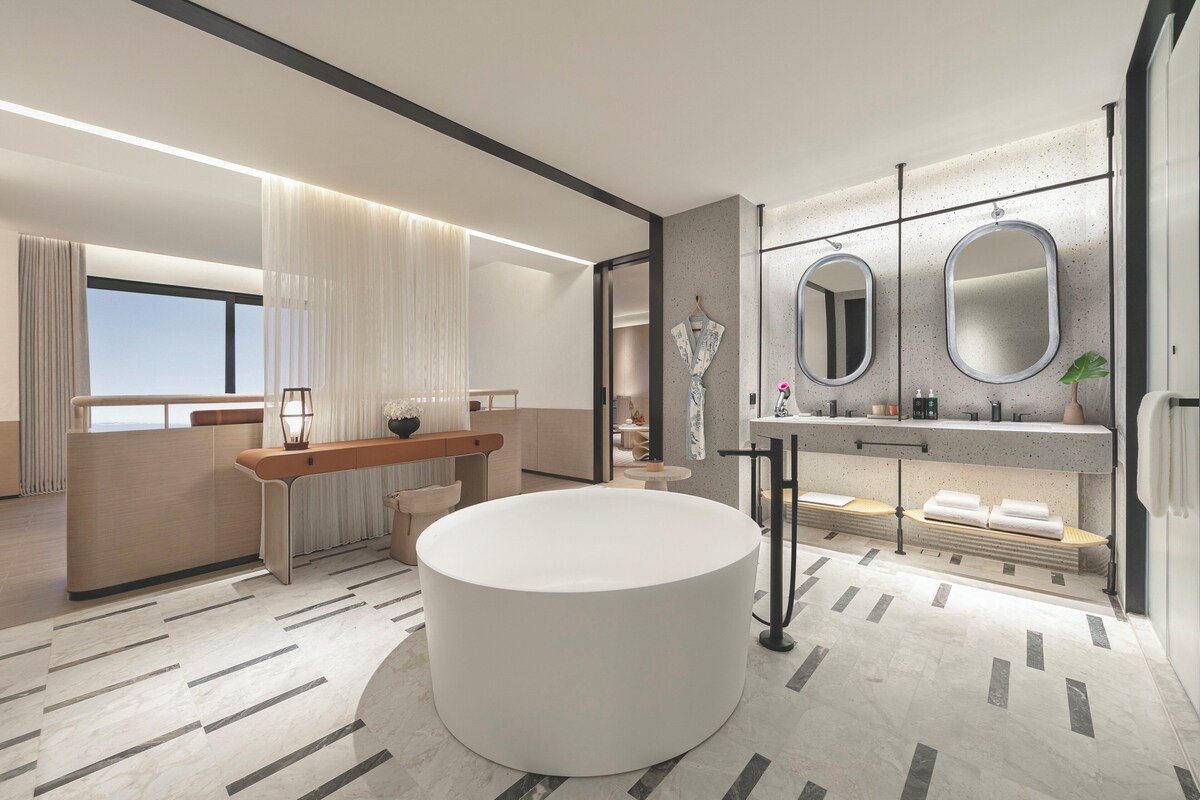 A modern bathroom with a large round bathtub, a vanity with two mirrors, and a seating area with a bench.
