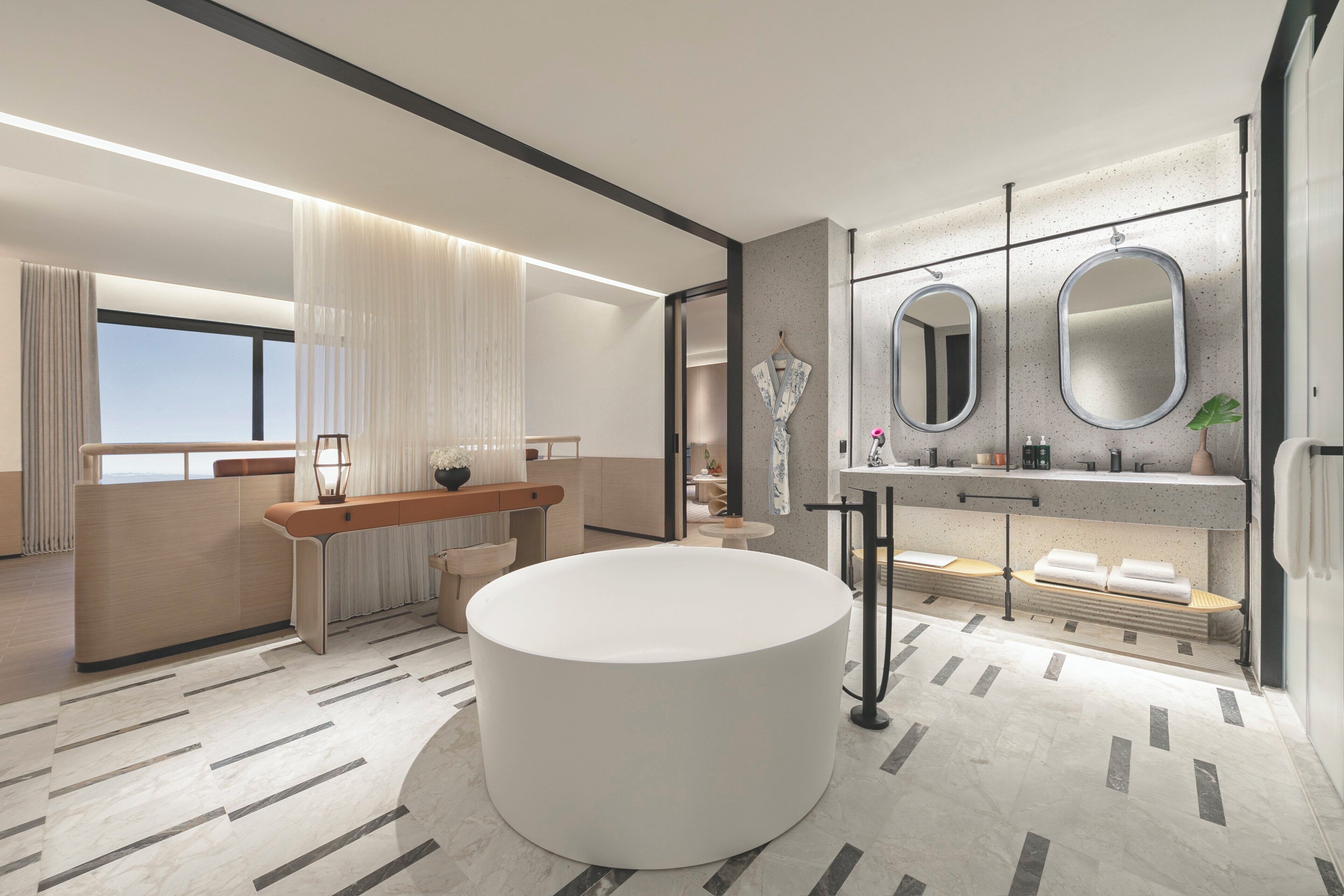 Een moderne badkamer met een groot rond bad, een wastafel met twee spiegels en een zithoek met een bank.
