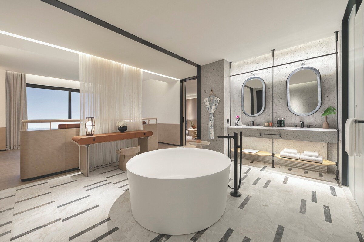 A modern bathroom with a large round bathtub, a vanity with two mirrors, and a seating area with a bench.