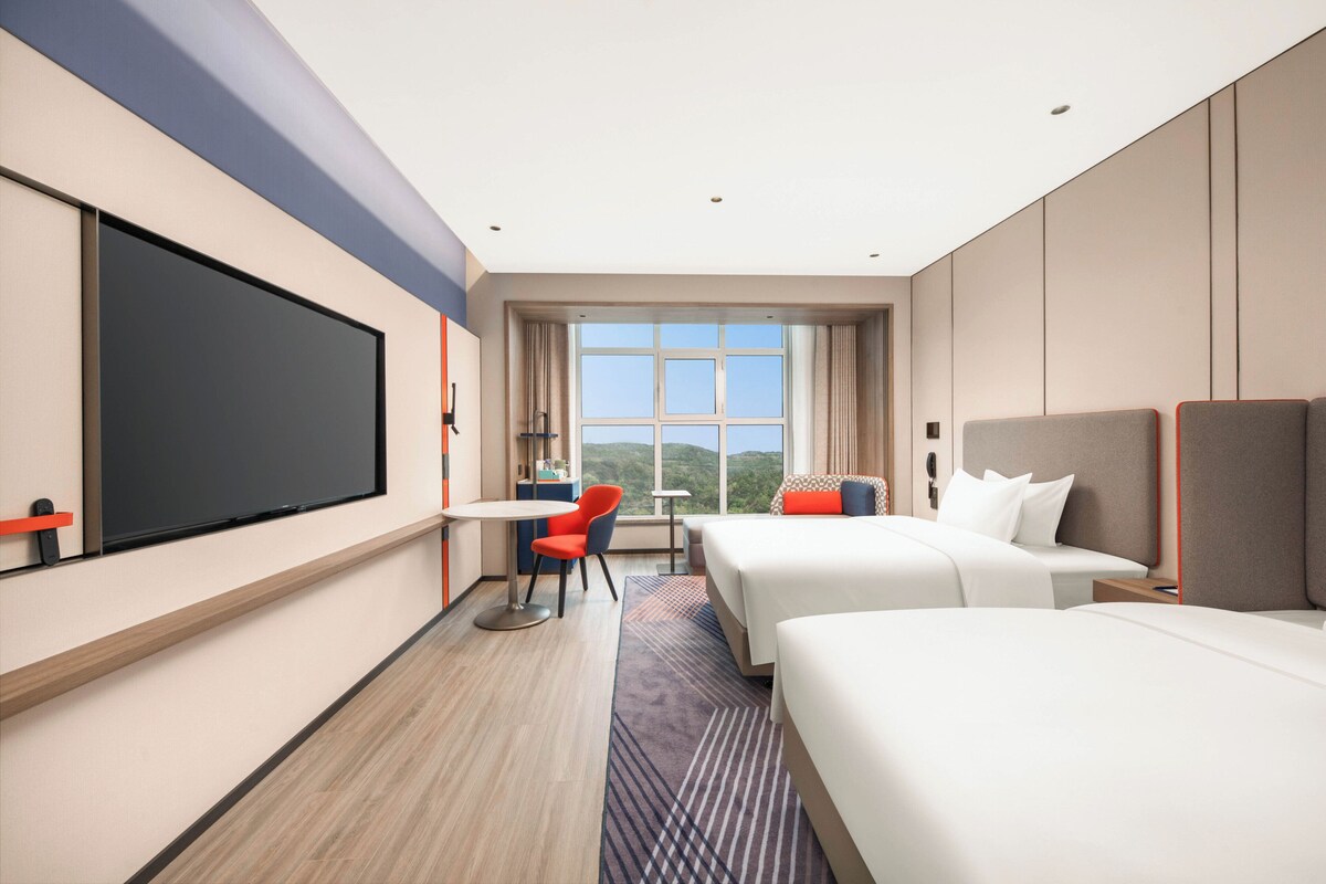 A hotel room with a large bed, a smaller bed, a desk, a chair, and a large window with a view of the mountains.