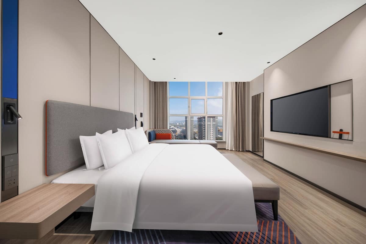 A modern hotel room with a large bed, a TV mounted on the wall, and a city view through the window.