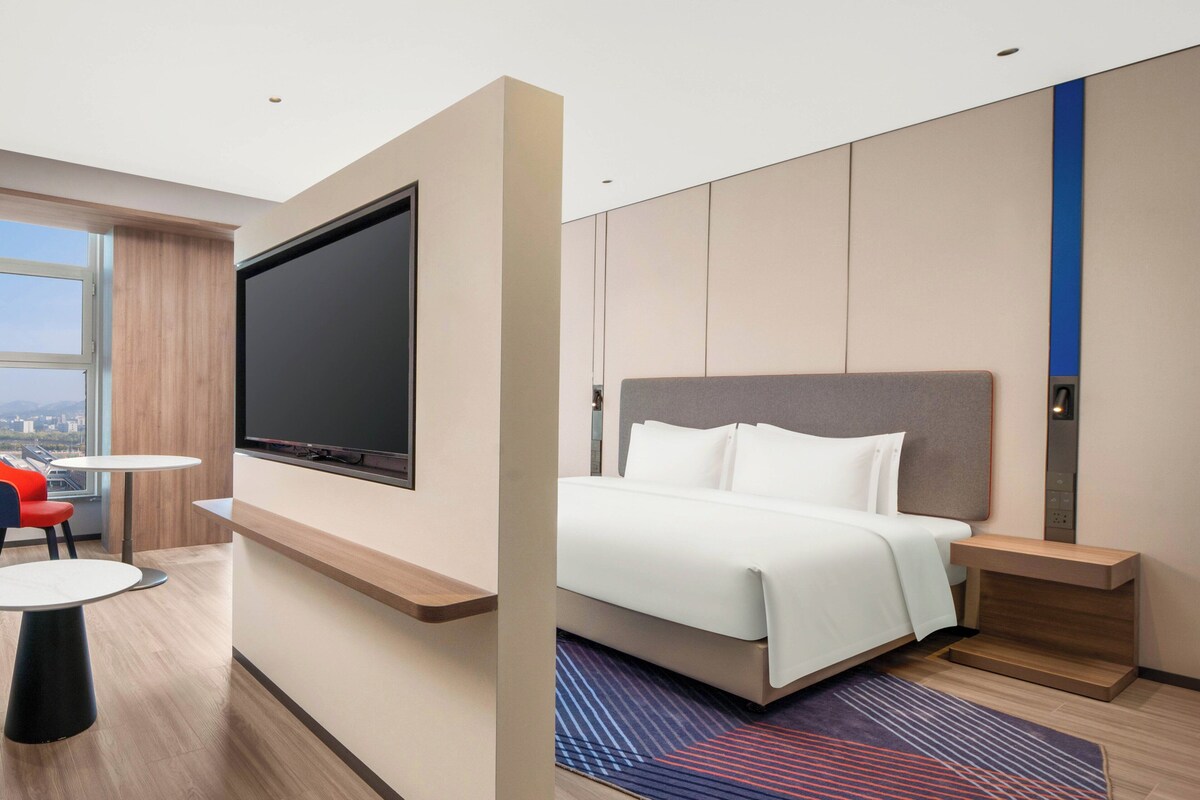 A modern hotel room with a large flat-screen TV, a bed with white linens, a small round table with a red chair, and a city view through the window.