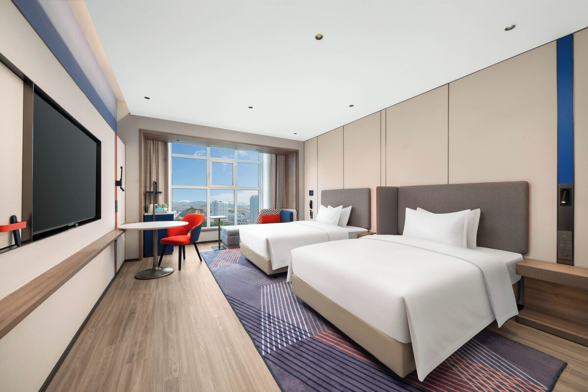 A hotel room with two beds, a desk, a chair, and a large window offering a city view.