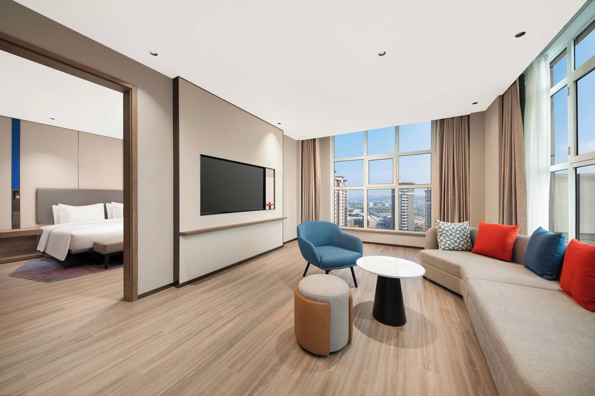 A modern hotel room with a large bed, a sofa, a blue armchair, a round coffee table, and a TV mounted on the wall.