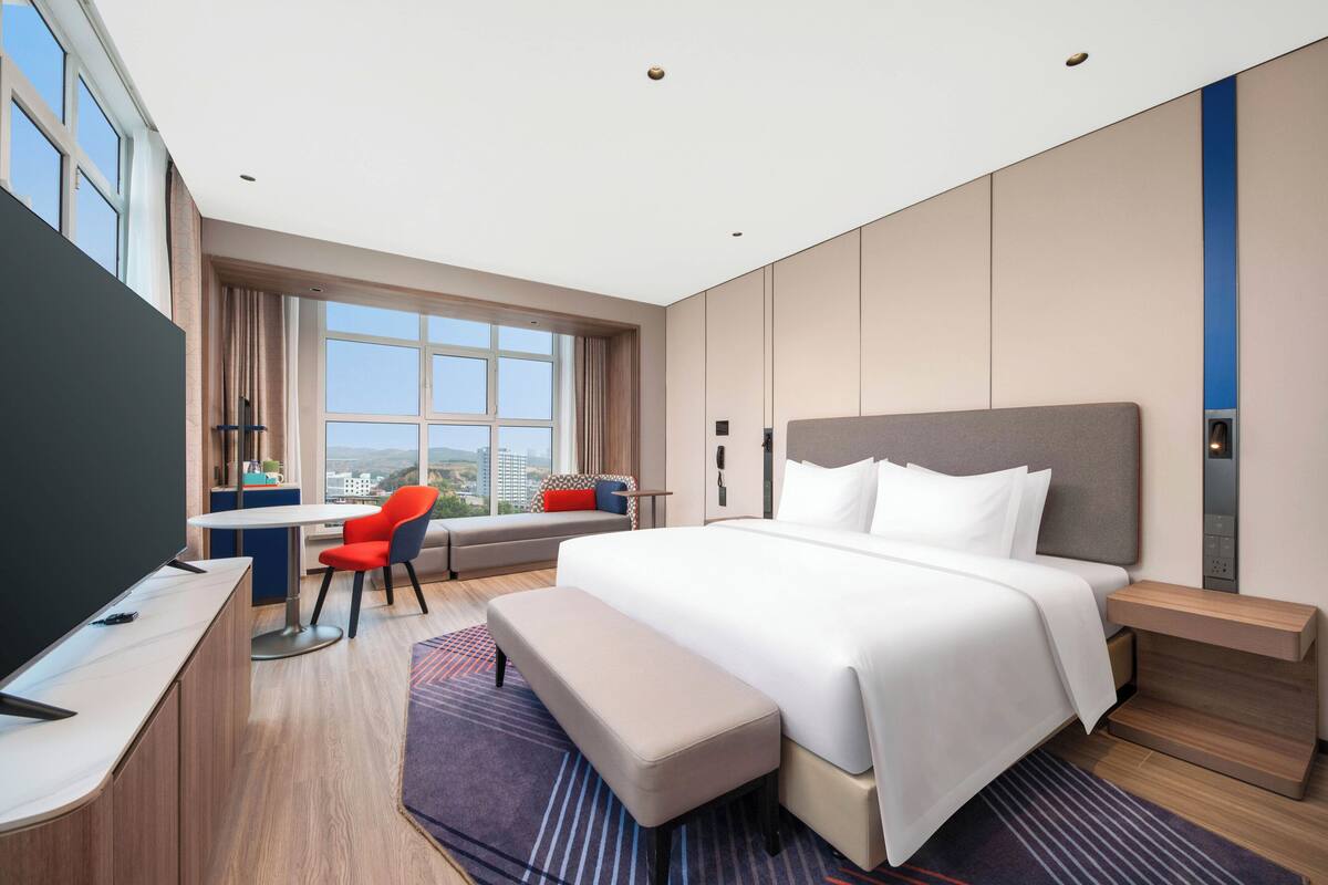 A modern hotel room with a large bed, a desk, a chair, and a view of the city.