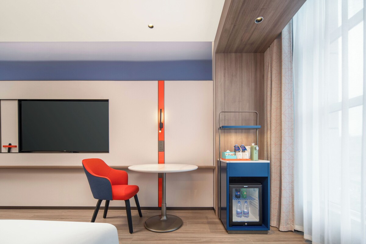 A hotel room with a bed, a TV, a small table with two chairs, a minibar, and a window with curtains.